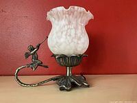 Front view of the lamp showing the white ruffled flower-shaped glass shade with mottled pattern and the metal base with fairy figure perched on the curved arm.