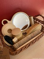Full view of the vintage French alabastrine dresser set items arranged in a wicker basket including a hand mirror, brushes, powder shaker and other grooming tools.