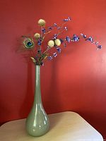 Green genie bottle decanter with dried floral arrangement in it, shown on beige surface against red wall