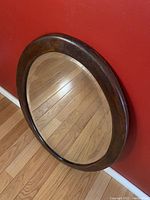 Photo showing the full view of the oval walnut framed beveled mirror resting against a red wall on a wooden floor, highlighting the shape and wood texture of the frame.