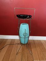 Full item view showing the turquoise textured glass cylinder in a standing metal frame with tripod legs and an ashtray dish on top against a red wall and wood floor.