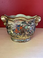 Front angle displaying detailed hand-painted scene of horsemen and mountainous landscape with curled gilt handles.