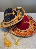 Two velvet Mexican sombreros, one black and one red, both decorated with detailed gold embroidery and gold braided chin tassels, heads together viewed from above and side.