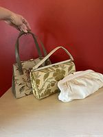 Front and profile view of three handbags: embroidered floral handbag, vintage Eatons floral clutch, white leather clamshell clutch on a flat surface against red background