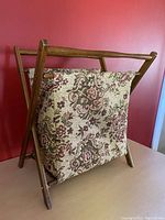 Front angle view of the vintage folding knitting bag showing the floral fabric container and wooden frame standing against a red wall.