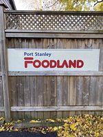 Front view of the large vintage Foodland sign mounted on a wooden fence outdoors, showing the entire sign with the text "Port Stanley FOODLAND" in blue and red.