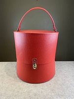 Front view of cylindrical red textured vinyl hat or wig carrying case with handle and clasp.