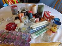 Various candles and candle holders on a table including small votive candles in packaging, pillar candles, jar candles, and decorative holders.