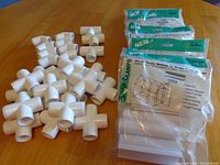 Photo showing loose white plastic pipe connectors of three different types (elbow, T, and cross) and five unopened 10 packs of white plastic snap clamps