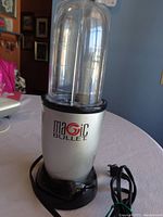 Front view of silver Magic Bullet blender base with attached tall clear plastic blending cup on top, sitting on a round table.