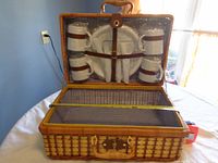 Open picnic basket showing secured plates, mugs, and utensils inside with blue and white checkered lining