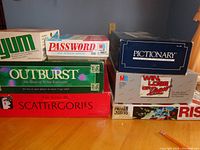Seven boxed board games stacked on a table showing box names Password, Outburst, Pictionary, Win Lose or Draw, Yum, Scatterories, Risk
