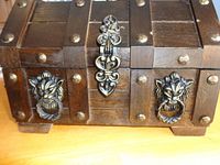 Close-up of front side of dark wooden chest showing its metal latch and decorative handles shaped like lion heads with rings.