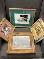 Four vintage framed artworks resting on a gray couch: one with a green mat and black and white photograph or print, one colorful floral painting in an ornate off-white frame, one impressionistic colorful small painting in a reddish frame, and one wood framed painting with figures and geese by a bare tree.