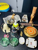 Top-down photo showing assorted ashtrays, bells, decorative wooden dagger, crystal block and other assorted items arranged on a black table.