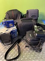 Photo displaying three camera bags and two digital cameras with cases and packaging on a metallic surface against a green background.