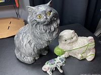 Photo of three cat statues together: large gray cat, white cat with green yarn, small floral-decorated cat