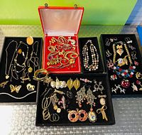Four trays on table showing assorted vintage and contemporary costume jewelry pieces including brooches, necklaces, bracelets, earrings.