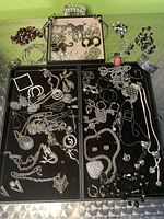 Full view of two black jewelry trays on silver table showing multiple necklaces, pendants, necklaces, bracelets and earrings