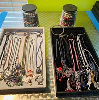 Photo of two trays with handmade necklaces on beige and black backgrounds with two large glass jars filled with jewelry behind them.