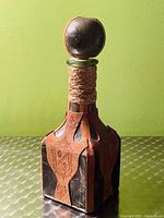 Front angled view of the square glass wine bottle wrapped in stitched leather with decorative emblem and twine wrapped neck topped with round leather-covered cork stopper.