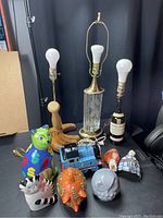 Photo showing three lamps: a wooden baseball glove and bat lamp measuring approximately 21 inches tall, an upcycled whiskey bottle lamp approximately 18.5 inches tall with a Caol Ila bottle base, and a Dresden-style lamp about 26 inches tall with a glass textured base and brass details. Also visible are five colorful money banks shaped in various figures and two small night lights.