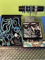 Photo showing multiple turquoise jewelry pieces including bead necklaces, bracelets, earrings in black trays and metal bracelet stands.