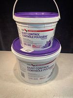 Two containers of CGC Sheetrock Dust Control drywall compound, stacked. Larger container at bottom with purple lid; smaller container on top with handle.