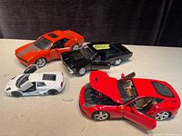 Top down view of four die cast metal cars showing the red Ferrari with open hood and doors, white Lamborghini with open scissor doors, black Dodge Charger with open driver's door, and orange 1966 Chevelle with open door.