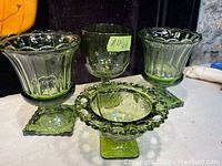 Four green glass items: three vases and one bowl. All have square pedestal bases and varying decorative rims. The bowl has a hobnail pattern around the scalloped rim. The vases have smooth to fluted edges with subtle vertical details.