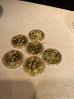 Six gold-colored physical Bitcoin commemorative coins arranged on a light surface. Three coins show the Bitcoin logo side, three show the circuit board side, all within clear plastic protective cases.