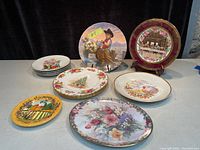 Photo showing a variety of plates including Christmas plates, floral design plates, and a Last Supper plate with burgundy border, all arranged on a table.