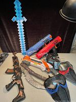 Toys including Nerf Rival guns in red, blue, white, a pixelated light-up sword, toy assault rifle, curved blade toy weapon, and two face masks arranged on a table.