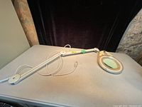Full view of an adjustable folding and extending magnifying lamp showing the arm, power cord, and lamp head with magnifying glass and circular light.