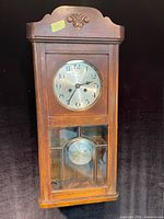 Full-frontal image of antique walnut wall clock showing carved top, metal clock face with Arabic numerals, two glass panels showing pendulum, wooden case with light wear.