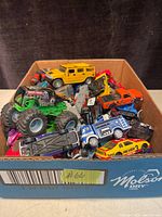 Box filled with assorted die-cast toy cars and trucks showing the top down view into the box with visible different colored vehicles including a yellow SUV and green monster truck.