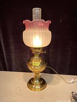 Brass table lamp with frosted pink ruffled art glass shade and clear glass chimney. Lamp is plugged in showing lit bulb inside frosted shade, demonstrating working condition.