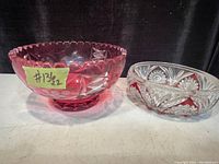 Two cranberry glass bowls placed on a surface, larger on left with scalloped rim and smaller on right with clear and cranberry glass cut pattern.