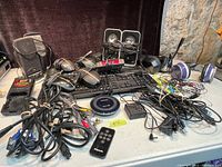 Wide view of electronics lot showing keyboards, mice, speakers, headphones, remote control, cables, and alarm clock.