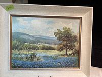 Framed signed landscape print in a light wooden frame with partly cloudy blue sky, trees, hills, and blue flowers in the foreground.
