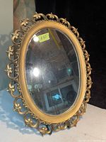 Oval wall mirror with gold ornate frame placed on a surface with reflections visible in the glass.