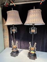 Pair of harlequin-style table lamps side by side on a surface against a dark background. Both lamps have decorative bases with harlequin figures, ribbed spheres, and crystal dangles beneath fabric shades.