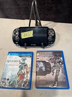 Sony PS Vita handheld console and two PS Vita game cases, one game identified as Assassin's Creed III: Liberation with visible front cover, the other game shows back cover with soldier illustration.