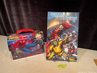 Photo showing the Transformers Volume 1 comic book standing upright with the Spider-Man metal lunch box in front, displaying both items clearly.