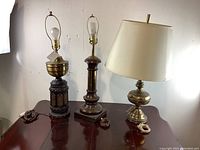 Photo of three assorted brass table lamps on a wooden surface against a plain wall.