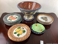 Photo showing entire ceramic dishes set arranged on a wooden surface including a large pedestal bowl and various plates with fruit and leaf designs.