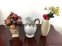 Image showing three decorative items on a wooden surface: a ceramic fruit arrangement, a white floral ceramic pitcher, and an off-white vase with artificial flowers.