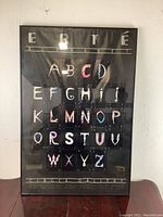Framed Erte Alphabet poster displayed vertically on a table against a wall, showcasing the letters arranged in a grid with a black background.