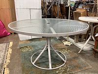 Round glass top outdoor patio table with white metal frame base shown from above and angled side view. Table has central hole for umbrella.