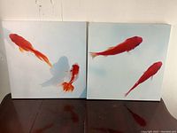 Pair of goldfish decorative plaques side by side on a dark wooden surface against a white wall.